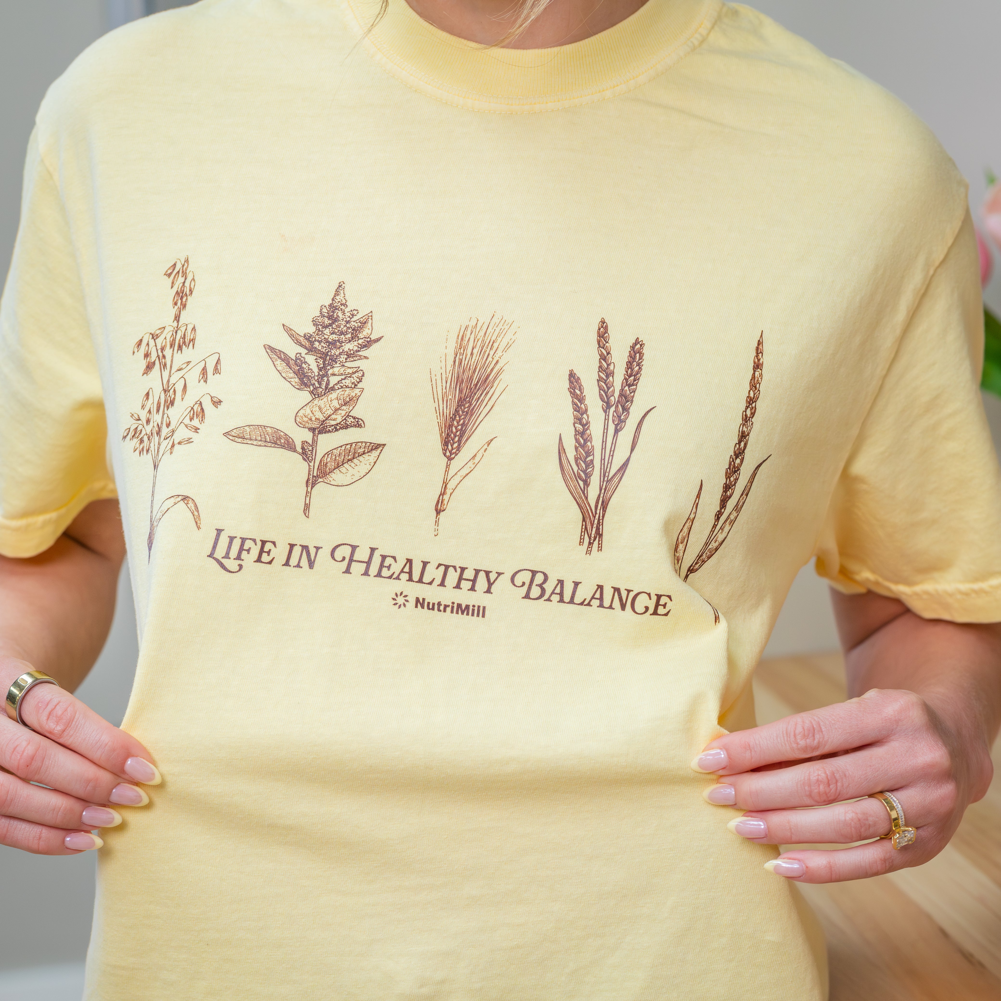 Close-up of a butter yellow NutriMill 'Life in Healthy Balance' t-shirt with brown wheat and grain illustrations, worn with hands at sides
