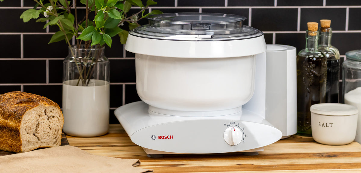 Bosch white universal plus mixer on a wooden counter next to bread, salt, oil and a plant with a black background