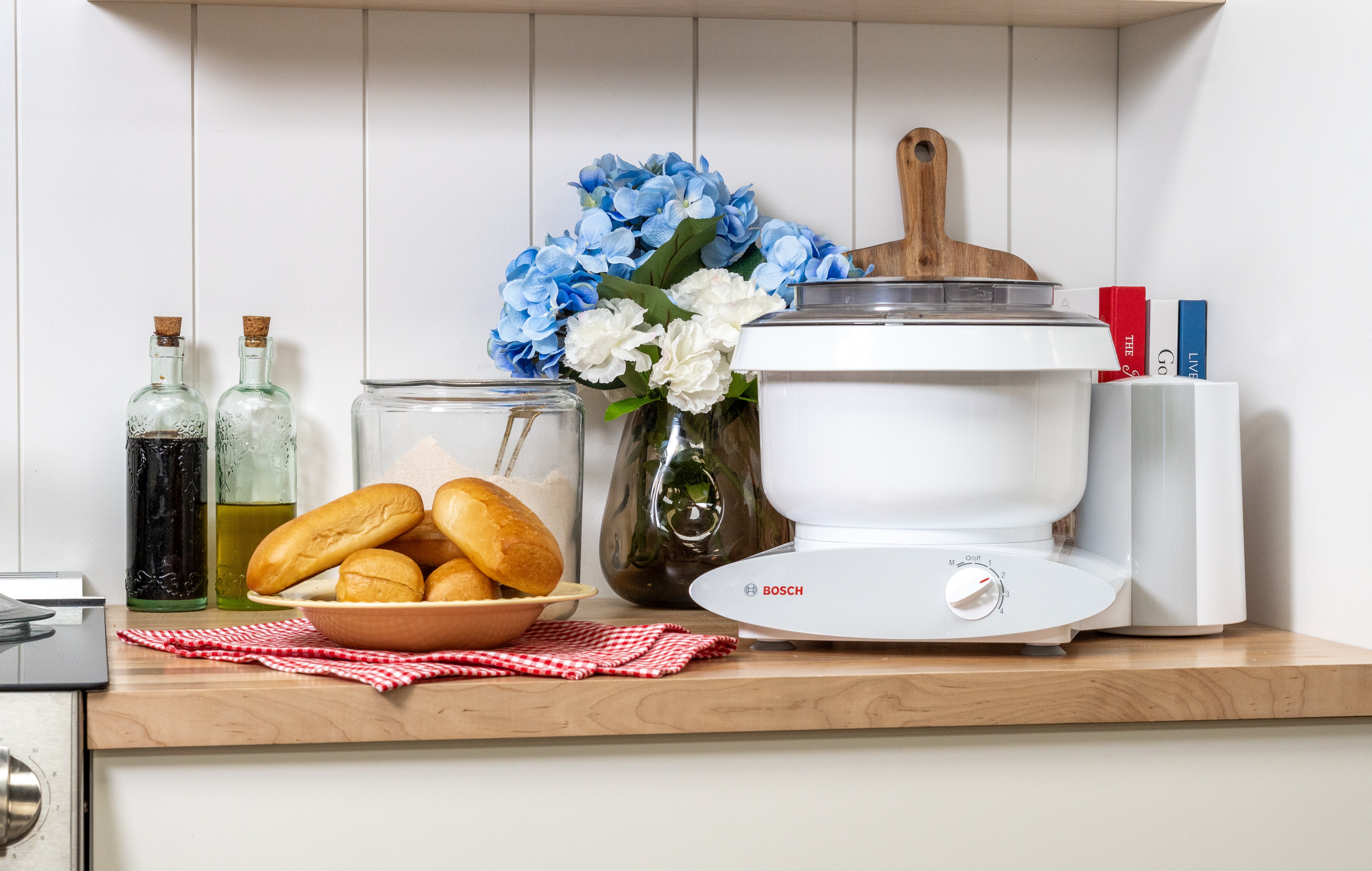 White Bosch Universal Plus mixer on a kitchen counter beside a tray of homemade buns and a plate of hot dogs