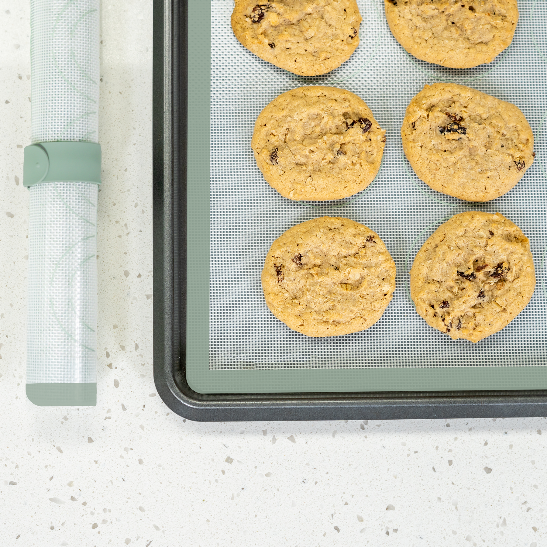 Top view of fresh-baked oatmeal raisin cookies on a NutriMill green silicone baking mat on a cookie sheet next to a rolled mat on a white speckled counter