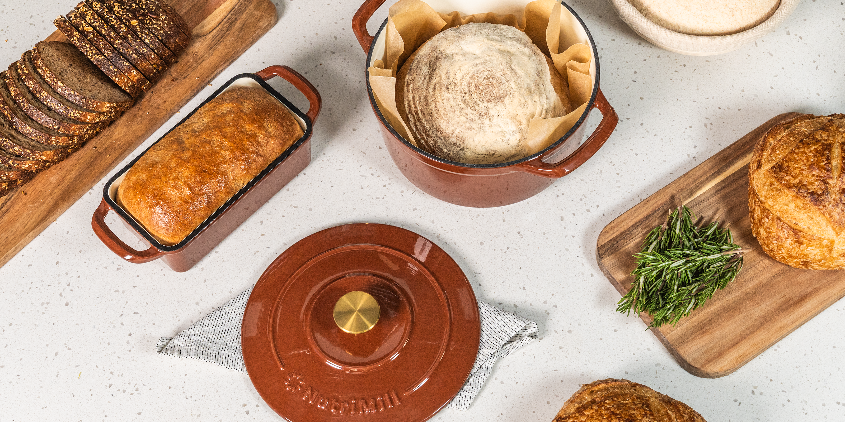 Dutch oven with sourdough inside and lid off next to bread pan with baked bread inside, next to sourdough with herbs and wheat bread on a white speckled counter.
