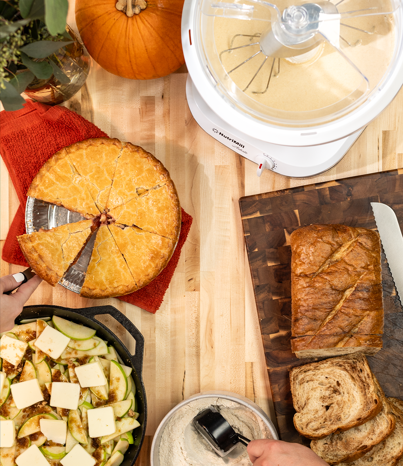 A pie, apple dessert, sliced loaf of bread, and a NutriMill Artiste mixer on wooden counter with fall decorations