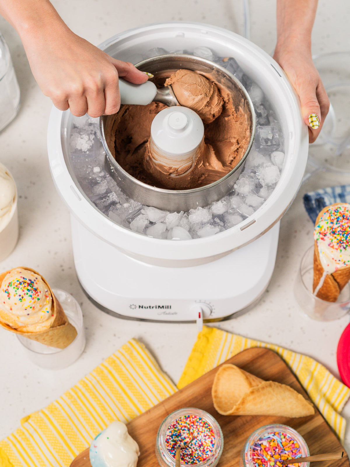 Hands scooping chocolate ice cream from the NutriMill Artiste ice cream maker attachment packed with ice, with waffle cones and sprinkles on the counter