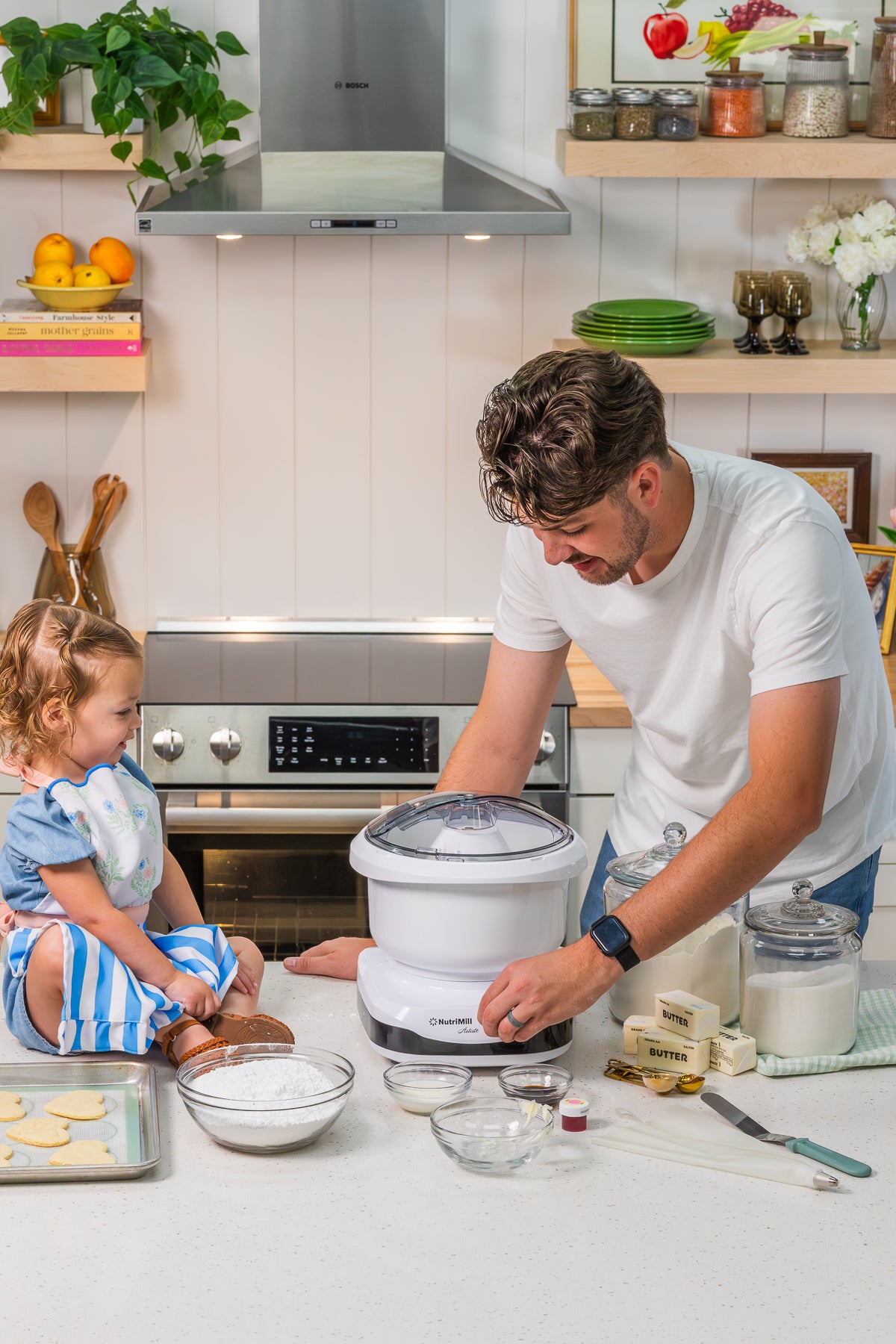 Man preparing ingredients at a white NutriMill Artiste mixer while a toddler girl sits on the counter beside bowls of baking ingredients