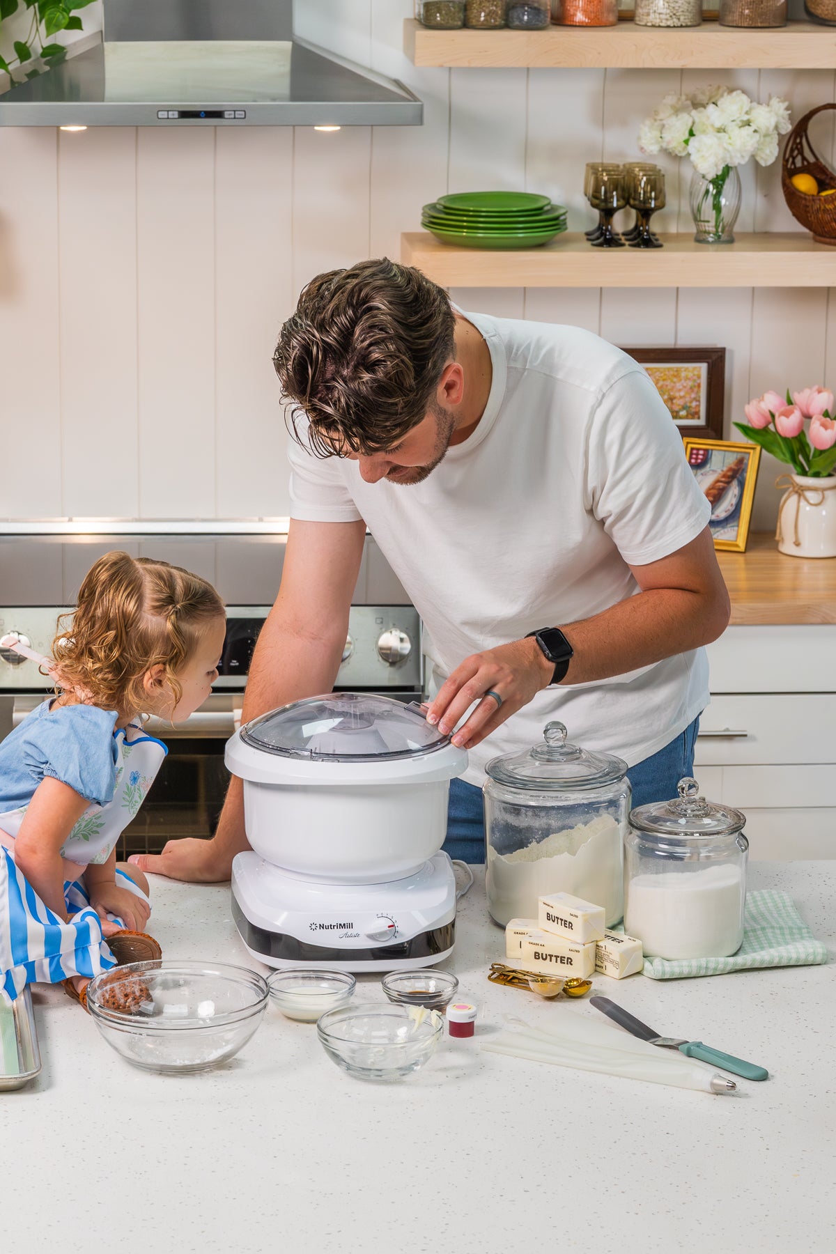 Man adding ingredients to a white NutriMill Artiste mixer with bowls of cocoa, flour, and butter while a toddler girl watches from beside the counter