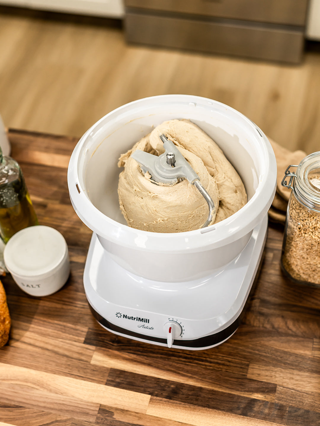 NutriMill artiste mixer mixing dough on a wooden counter next to salt, grains, and oil