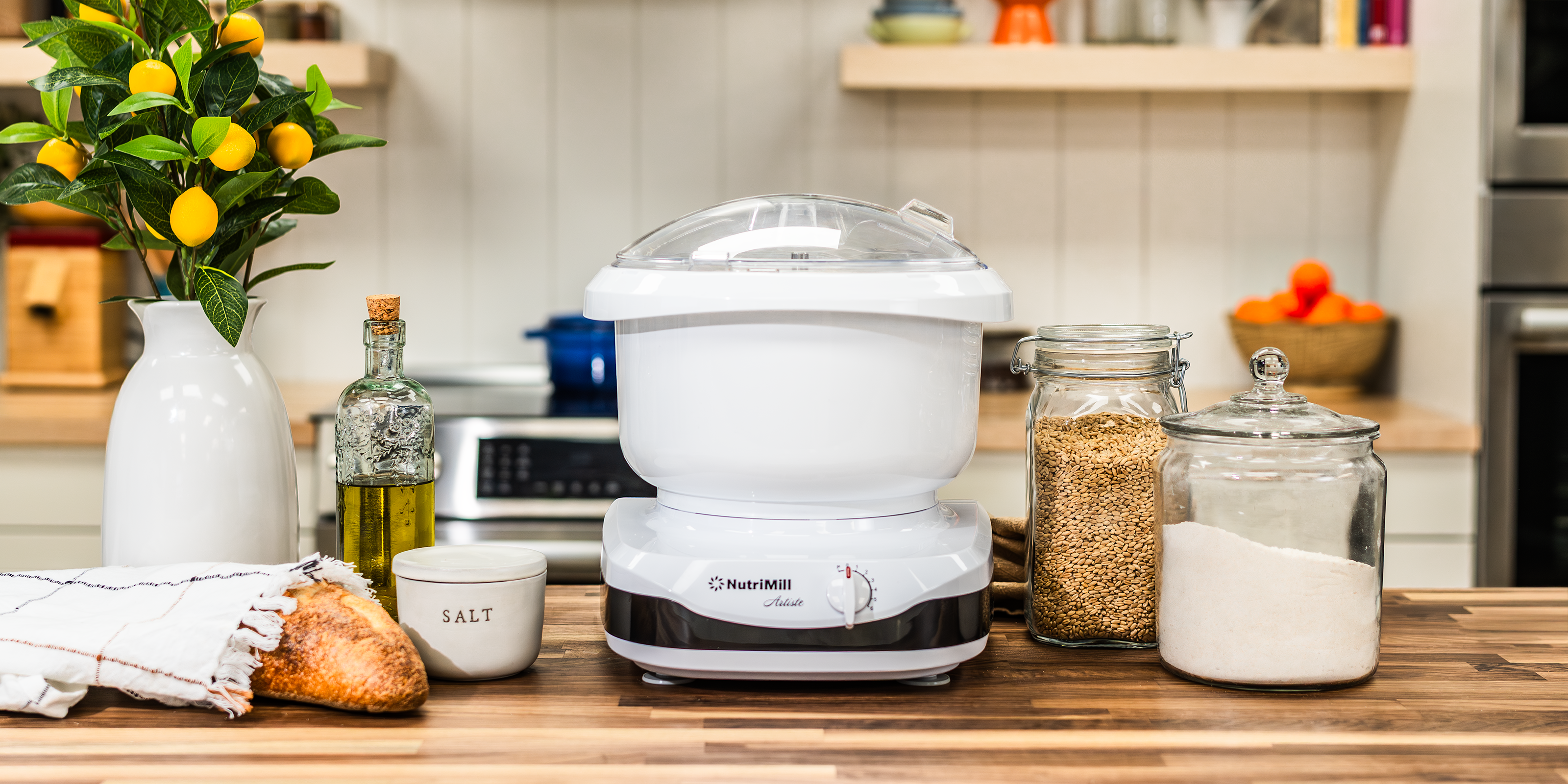 NutriMill artiste mixer on a wooden kitchen counter next to bread and baking ingredients