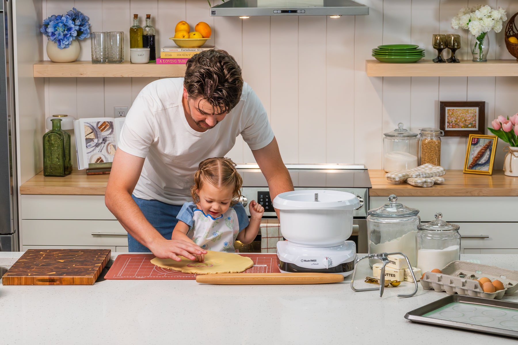 Man helping a toddler girl roll out sugar cookie dough with a rolling pin on a NutriMill pastry mat next to a white NutriMill Artiste mixer, butter, and eggs
