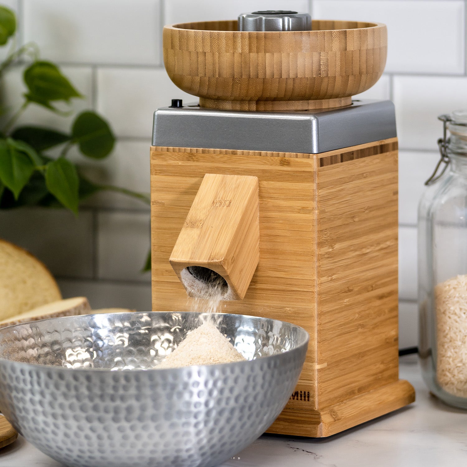 A NutriMill harvest grain mill with a silver trim milling grains into a silver bowl with a white wall in the background