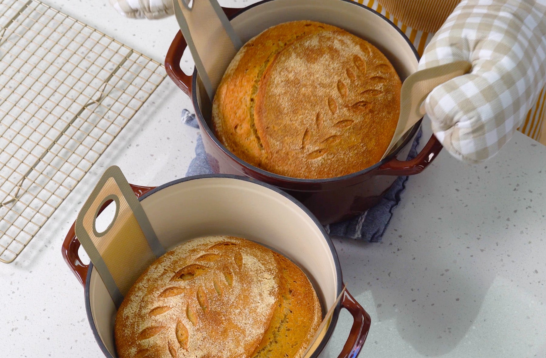 Two sourdough boules with leaf scoring patterns being lifted out of red enameled Dutch ovens with oven mitts on a white speckled counter