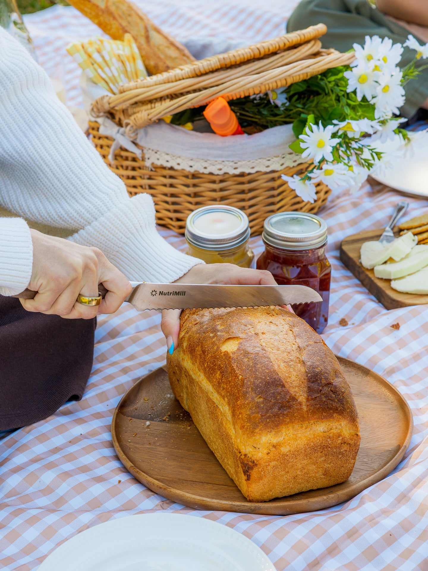 Person slicing a loaf of homemade bread at a picnic with a basket and jam jars