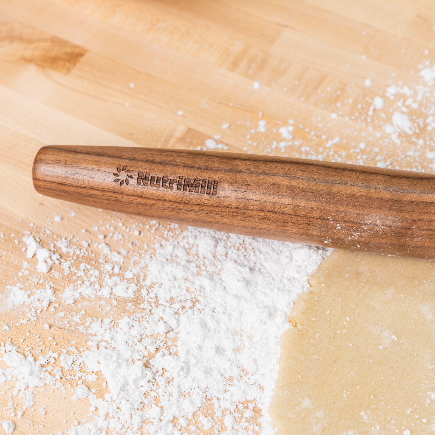 Close-up of the engraved NutriMill logo on the walnut French rolling pin, resting on a light wood board dusted with flour