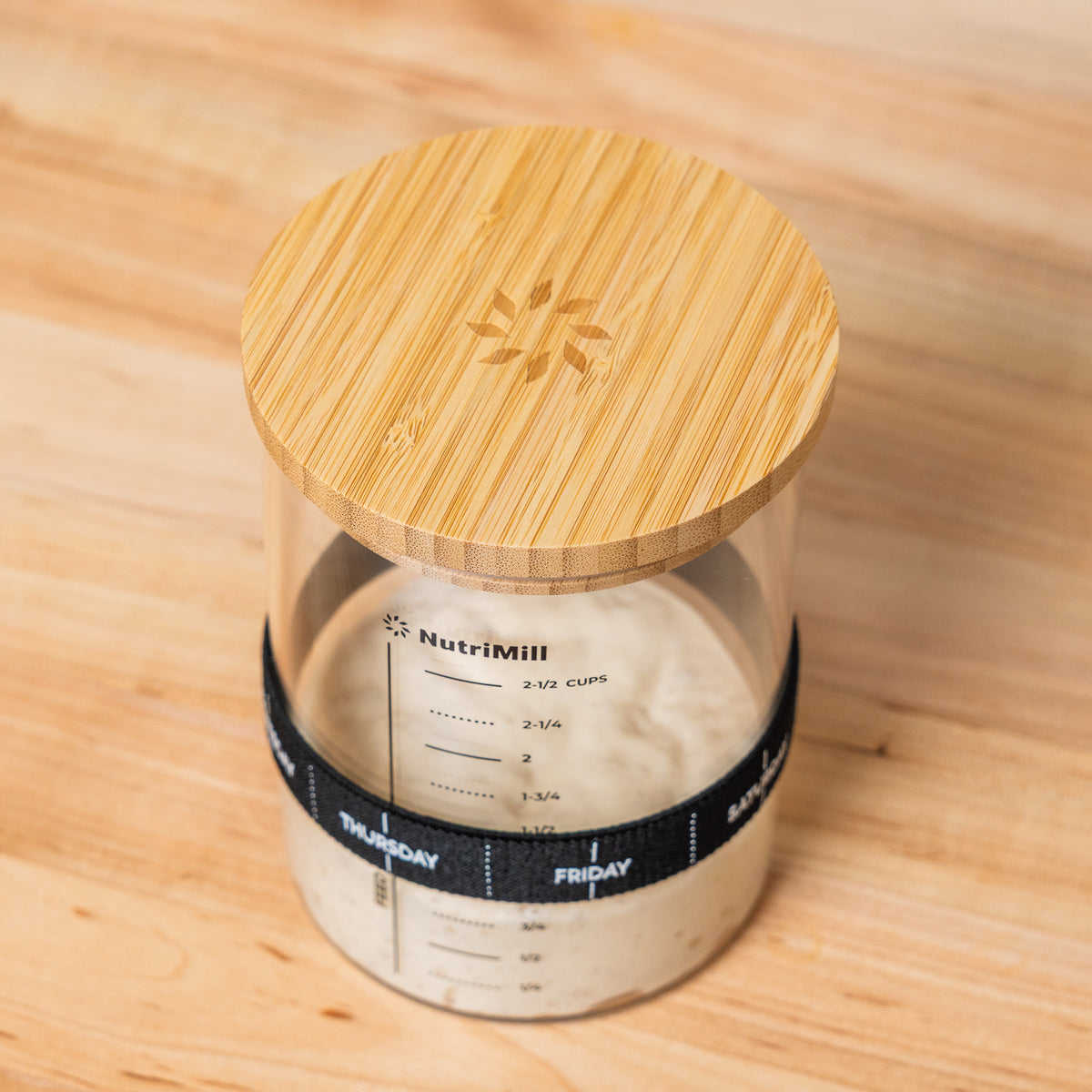 Close-up angled view of the NutriMill sourdough starter jar's bamboo lid with embossed NutriMill logo, the jar below filled with starter and wearing a day-of-the-week feeding band