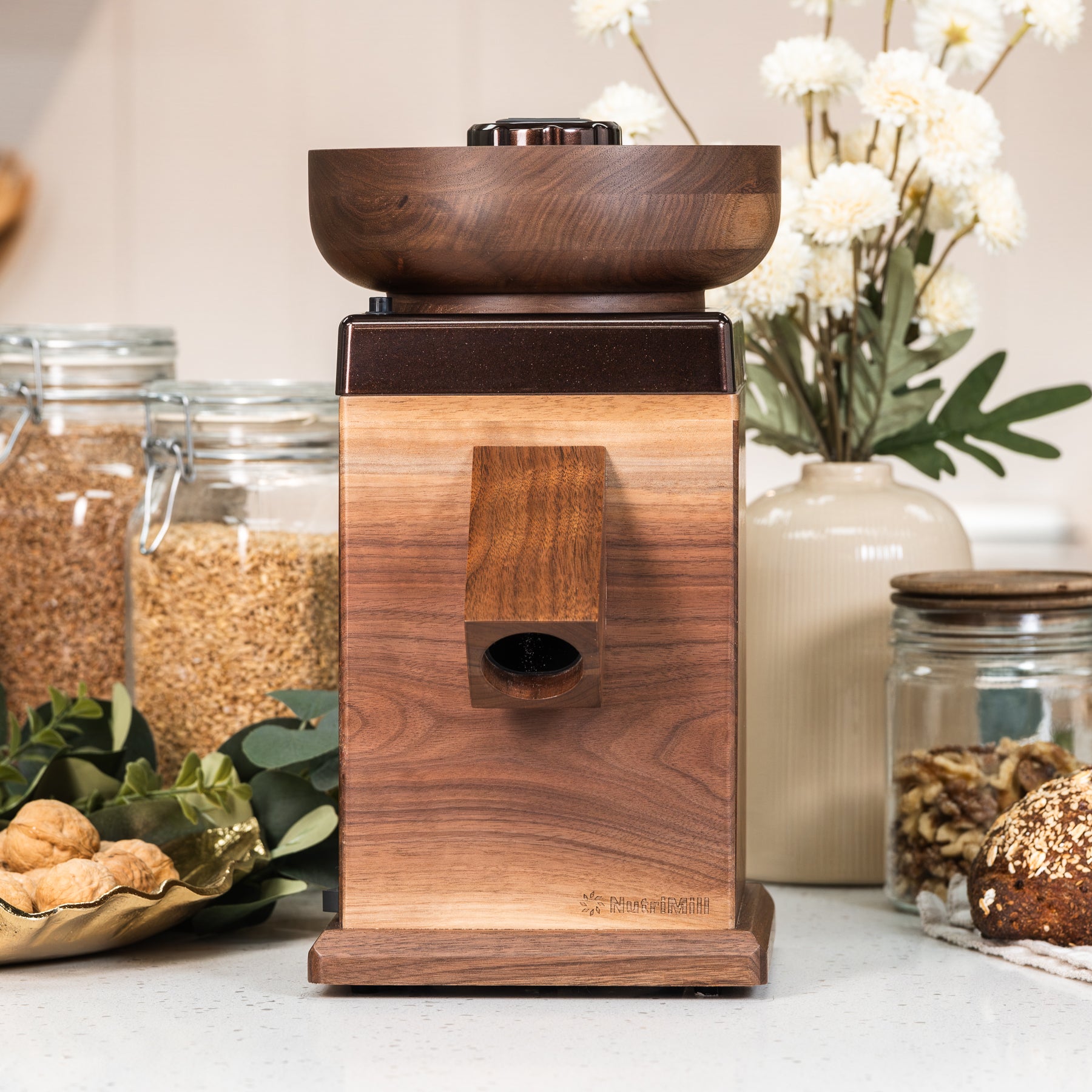 NutriMill harvest Walnut grain mill with cocoa-colored trim on white counter with plants and grains in background