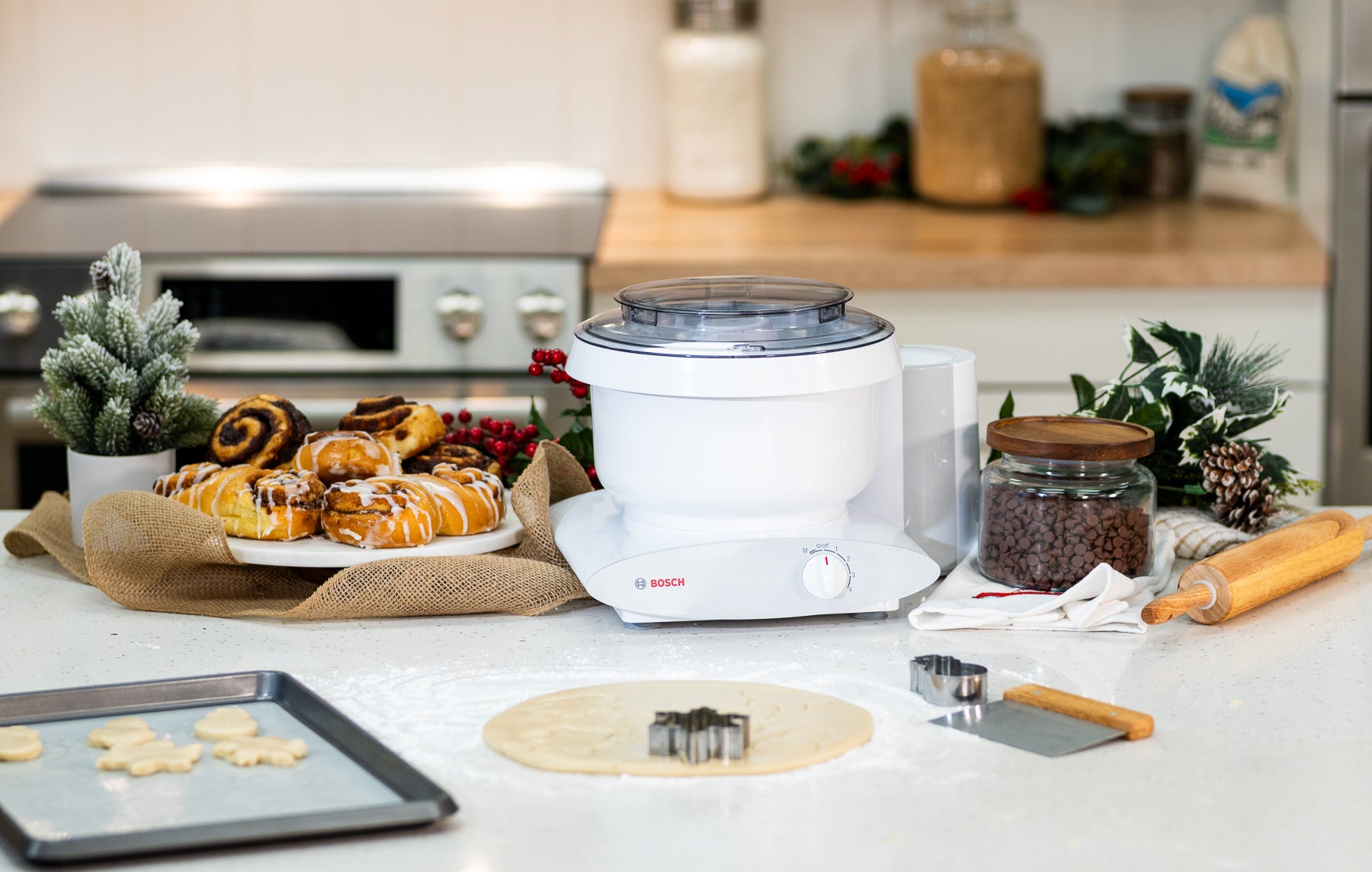 White Bosch universal plus mixer on white counter next to a plate of cinnamon rolls, Christmas decorations, dough, dough tools, and a jar of chocolate chips
