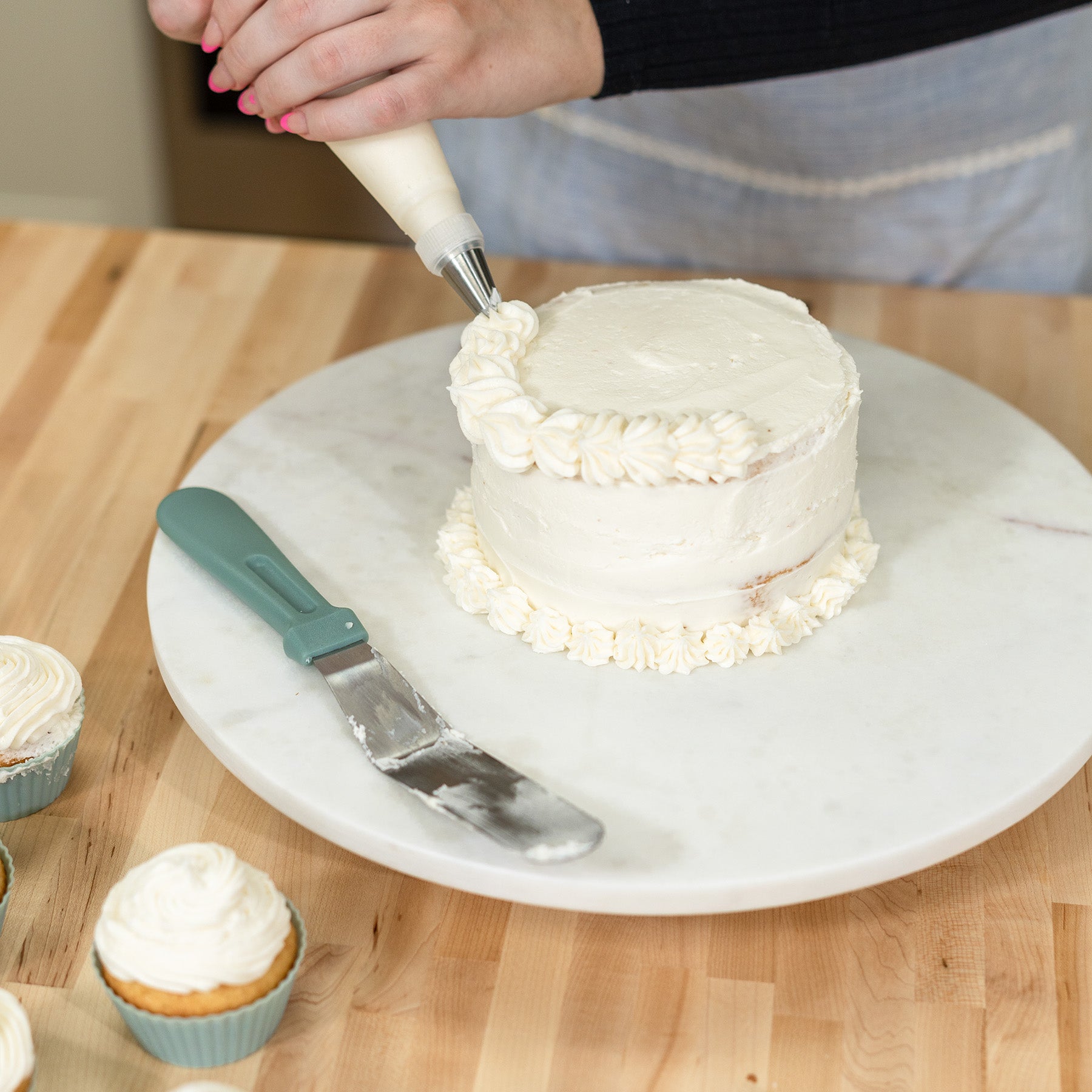 A woman icing a white cake with the NutriMill icing kit on a white platter sitting on a wooden counter