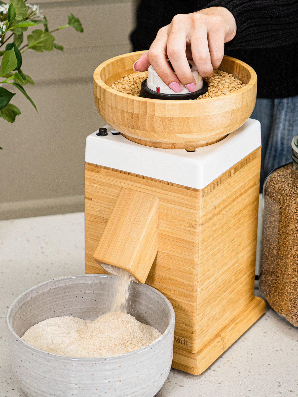 A woman milling flour with a NutriMill harvest grain mill with a white trim on a white kitchen counter